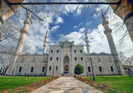 Fotografta Parcali Bulutlu Bir Gokyuzu Altinda Agaclar Ve Cimenlik Alanla Cevrili Dort Minareli Bir Cami Goruluyor Fotografta Parcali Bulutlu Bir Gokyuzu Altinda Agaclar Ve Cimenlik Alanla Cevrili Dort Minareli Bir Cami Goruluyor