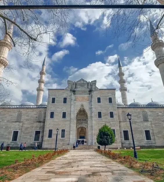 Fotografta Parcali Bulutlu Bir Gokyuzu Altinda Agaclar Ve Cimenlik Alanla Cevrili Dort Minareli Bir Cami Goruluyor