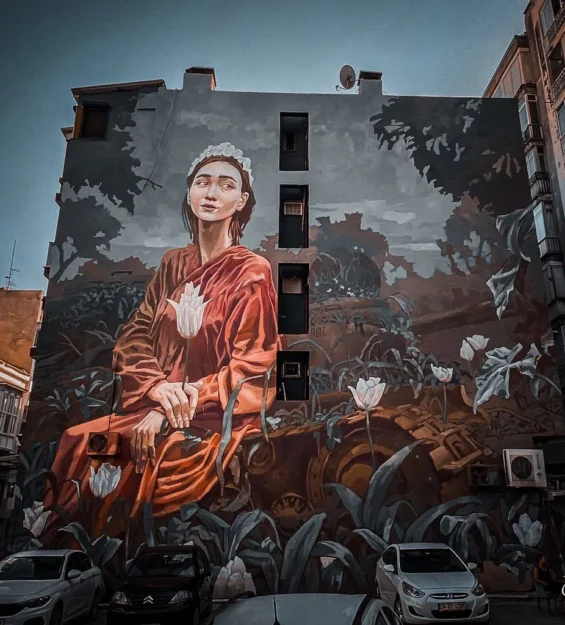 Çiçekler ve paslı bir tankın ortasında kırmızı elbiseli bir kadının yer aldığı büyük duvar resmi. Kentsel ortamda sokak sanatı. by fotoloji