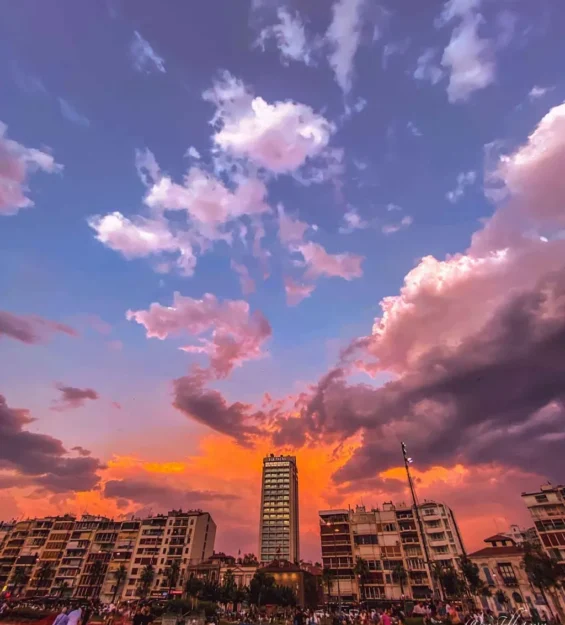 Deniz kıyısındaki binaların üzerinde pembe ve turuncu bulutlarla gün batımı manzarası. by fotoloji