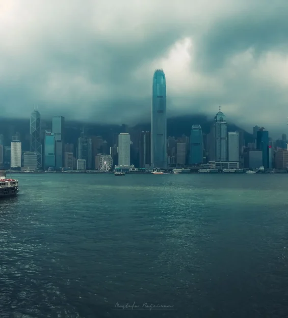 Limandan görülen Hong Kong silüeti, bulutlu bir gökyüzünün altında ikonik gökdelenleri sergiliyor. Ön planda yolcu gemisi. _by fotoloji
