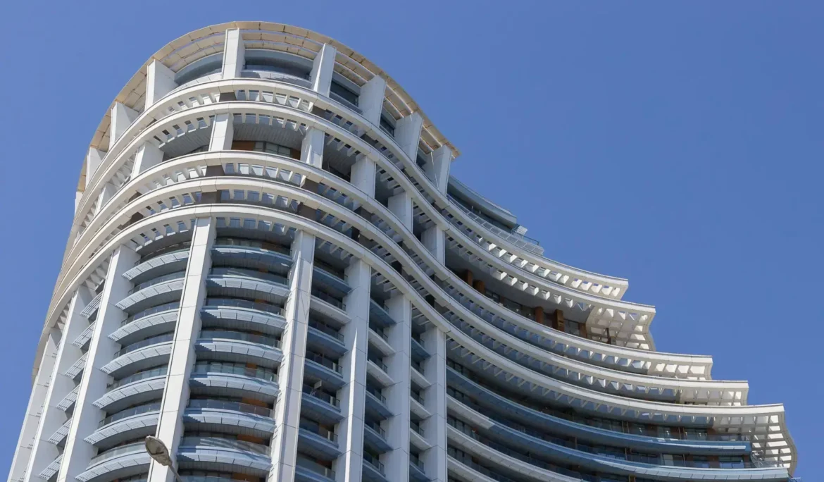 Açık mavi gökyüzüne karşı balkonlu ve mavi vurgulu, kavisli modern bina.