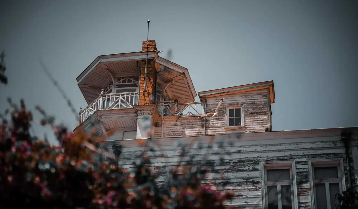 Kuleli ve balkonlu, kısmen yeşilliklerle örtülü, çürümeye yüz tutmuş ahşap bina.