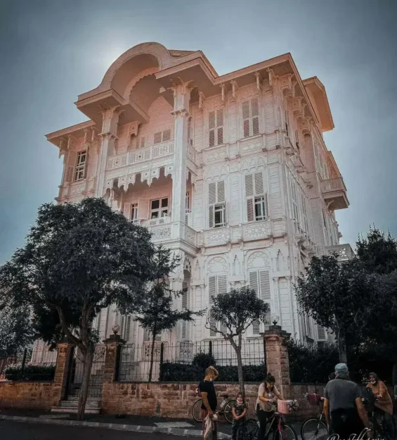 Ağaçlar ve bisikletli insanlarla çevrili, karmaşık detaylara sahip büyük beyaz Osmanlı dönemi konağı.