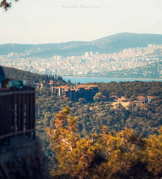 Türkiye'nin İzmir şehrinin panoramik manzarası, kısmen yıkık bir bina ve seyir terasında oturan izleyicilerin yer aldığı manzaralı bir yerden.