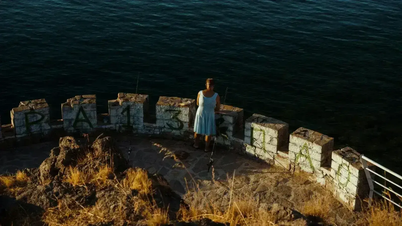 Deniz kenarında balık tutan kadın, grafiti bulunan taş duvarın üzerinde duruyor.