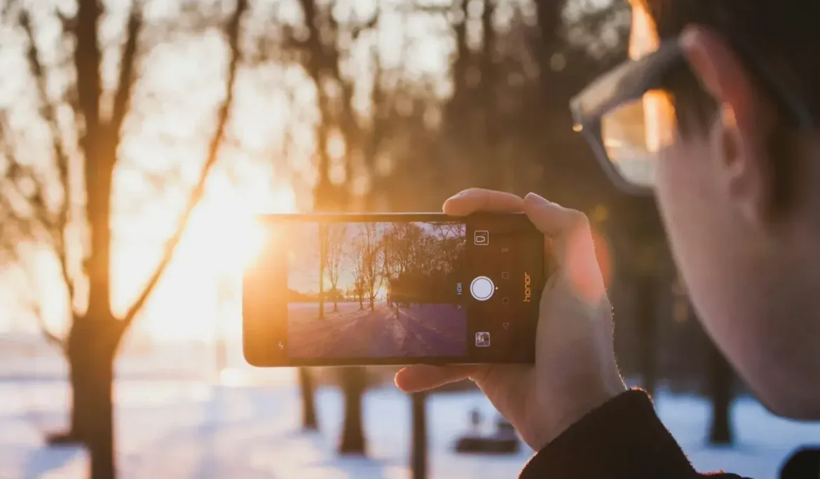 Gün batımında karlı bir kış manzarasını fotoğraflamak için telefon kamerası kullanan kişi.