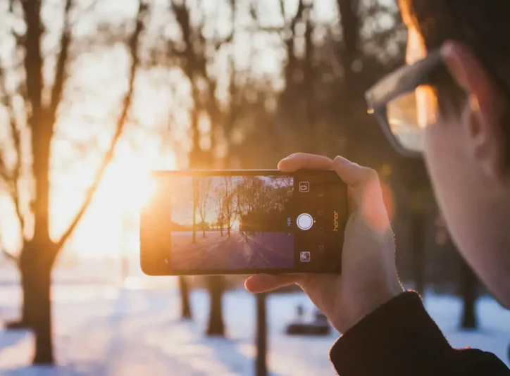 Gün batımında karlı bir kış manzarasını fotoğraflamak için telefon kamerası kullanan kişi.