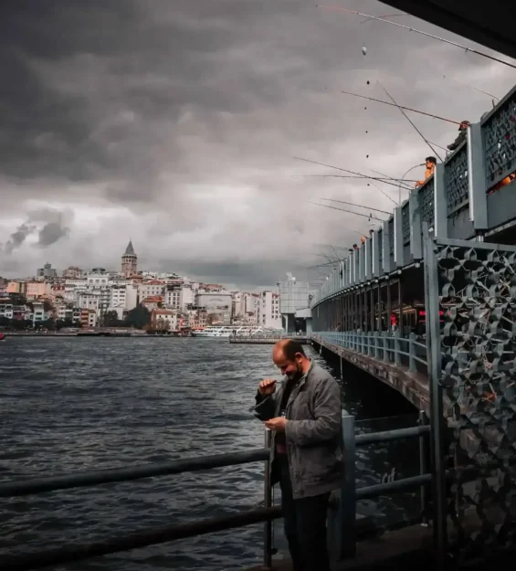 İstanbul köprüsünde telefona bakan adam, bulutlu gökyüzü, arka planda Galata Kulesi.