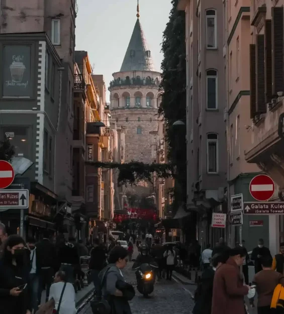Dar bir İstanbul sokağından Galata Kulesi manzarası, yürüyen insanlar ve bir motosiklet.