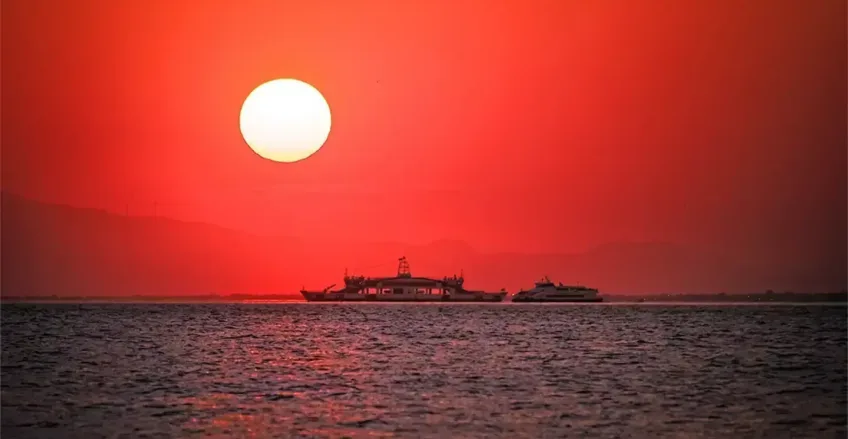 Sakin suyun üzerinde canlı kırmızı gün batımının önünde silüet oluşturan feribotlar.