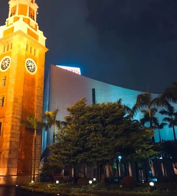Arka planda modern binalar ve palmiye ağaçları bulunan, aydınlatılmış Hong Kong Saat Kulesi'nin gece görünümü.