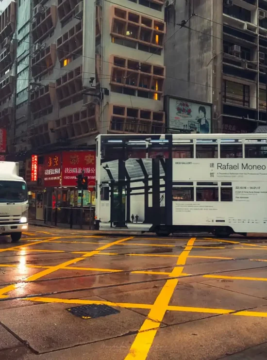 Rafael Moneo'nun reklamını yapan tramvay ve bir kamyonun yer aldığı yağmurlu Hong Kong sokak manzarası.