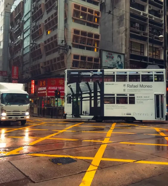 Rafael Moneo'nun reklamını yapan tramvay ve bir kamyonun yer aldığı yağmurlu Hong Kong sokak manzarası.