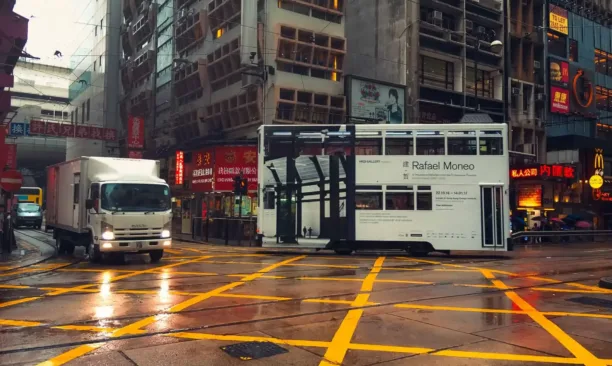 Rafael Moneo'nun reklamını yapan tramvay ve bir kamyonun yer aldığı yağmurlu Hong Kong sokak manzarası.