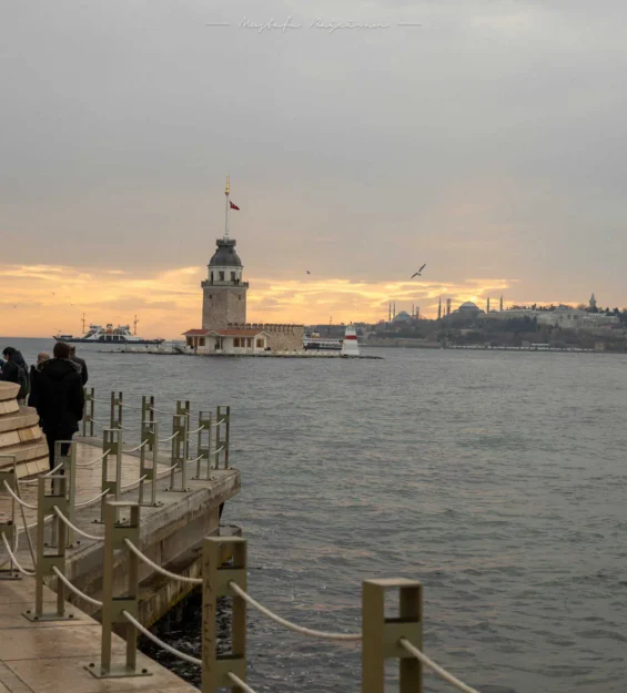 Kız Kulesi'nin İstanbul gün batımı manzarası ve sahil şeridinde yürüyen insanlar.