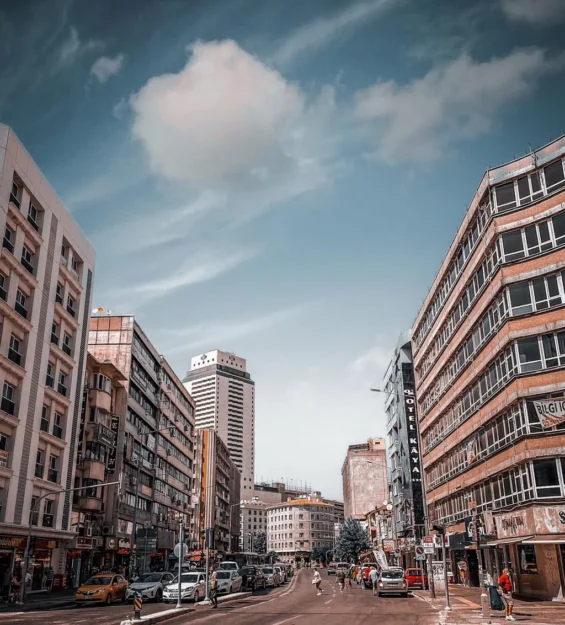 Türkiye'de parçalı bulutlu gökyüzünün altında binalar ve park edilmiş arabalarla şehir sokak manzarası.