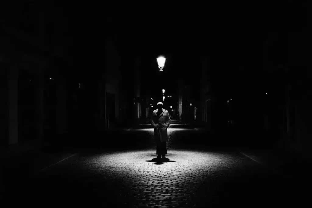 Karanlık sokakta ışık altında duran trençkotlu adam. Siyah beyaz fotoğrafçılık örneği.